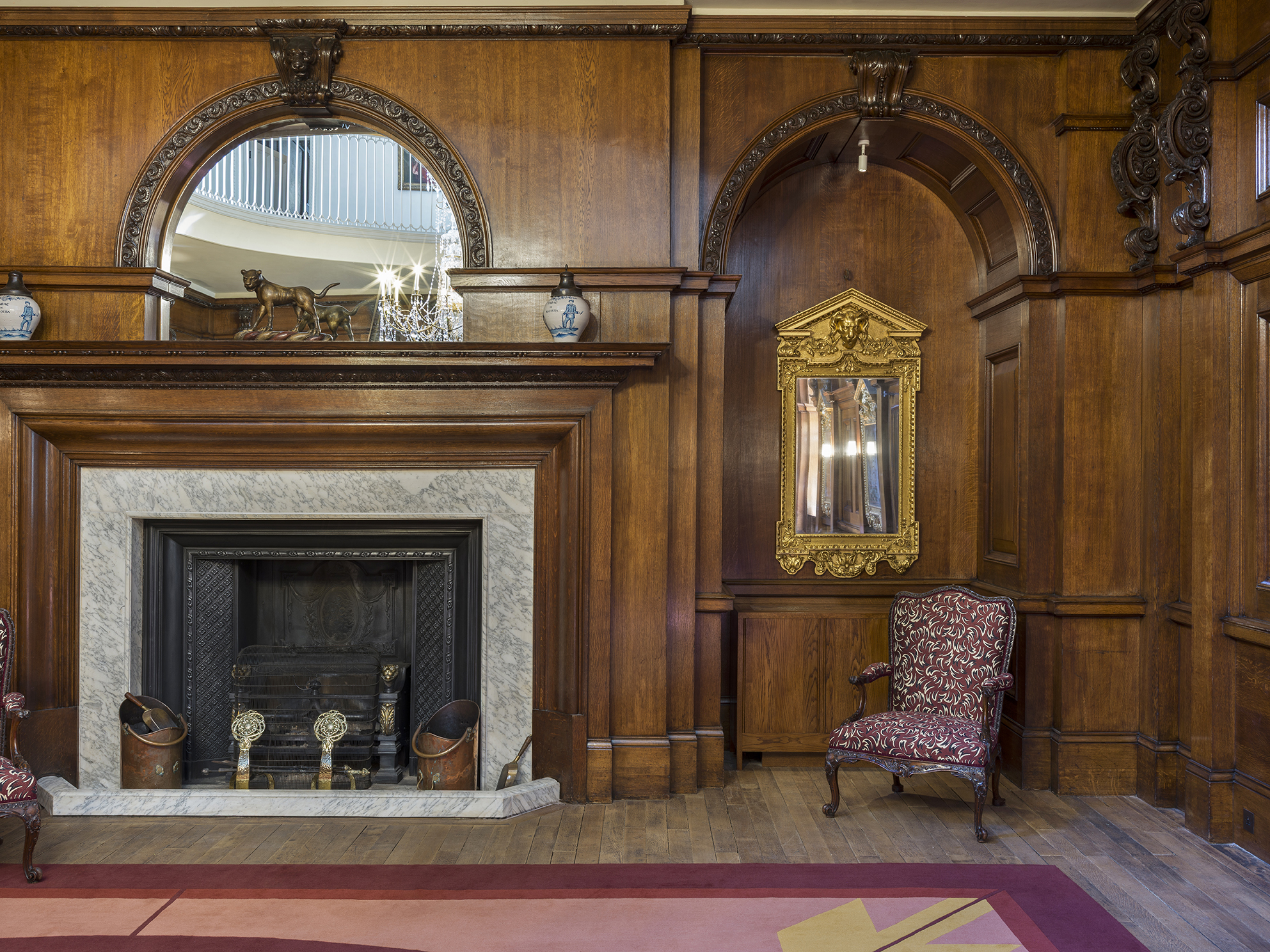 The Outer Hall, connected to the Great Hall through glazed doors, serves as a two-level reception area. The original timber flooring has been uncovered and a custom rug from The Rug Company, inspired by the Brangwyn paintings' colours in the adjacent hall, adorns the floor. This rug features a radial pattern that echoes the oculus on the gallery level above. A key feature of the room is the Malmsbury Chandelier, which dramatically spans both levels through the oculus. The upper level's ornately corniced walls and ceiling have been refreshed with a neutral colour palette, enhancing the prominence of the large gilt-framed portraits that line the walls. Additionally, original glazed doors provide access from the Outer Hall Gallery to a newly refurbished terrace outside.